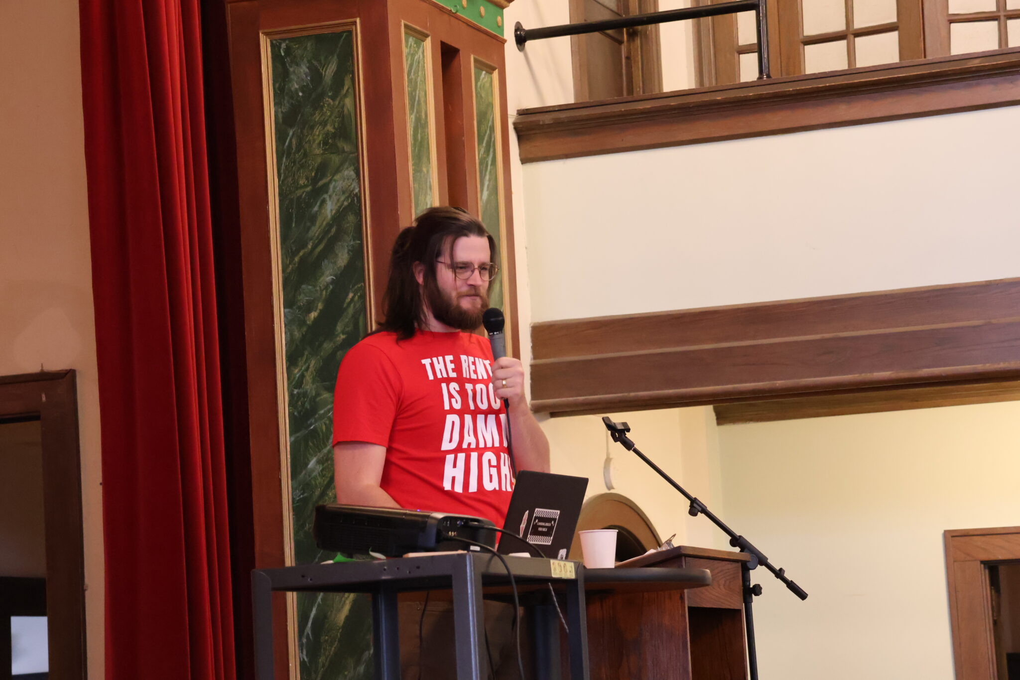 IMG_8825-2048×1365 A tall man with long brown hair wearing a red Rent is Too Damn High shirt and speaking into a microphone.