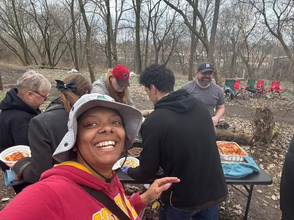 Khadja wearing a gray hat and red sweatshirt, smiling in front of people filling plates of food at Fallen Angels encampment